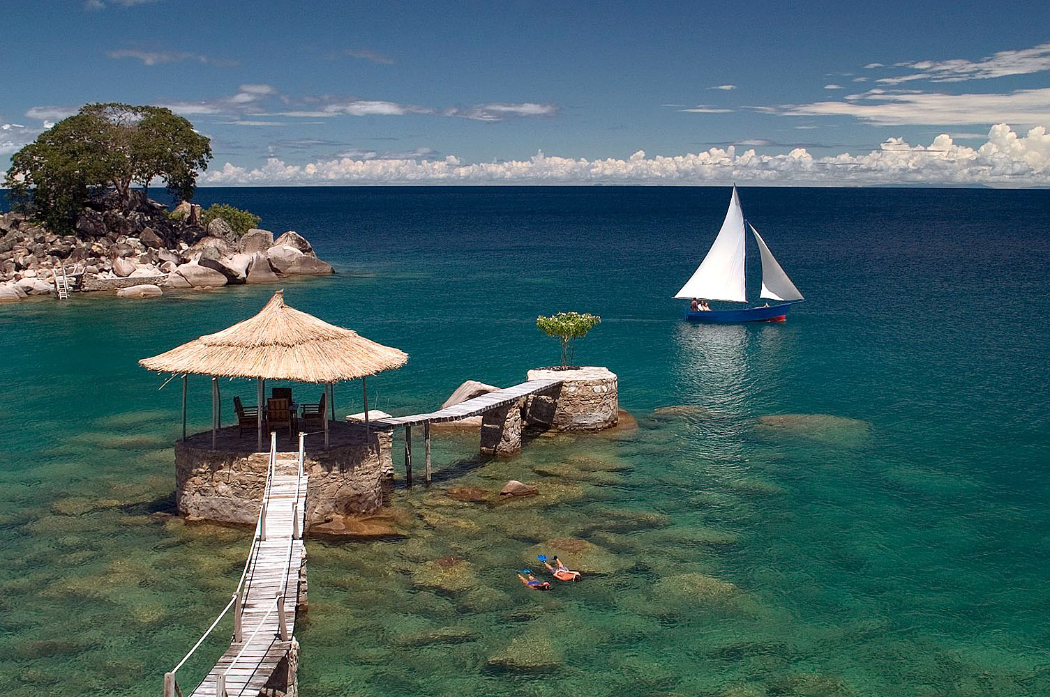 Traditionelles Segelboot auf dem türkisblauen Malawisee vor einer kleinen tropischen Insel mit Steg und Pavillon.