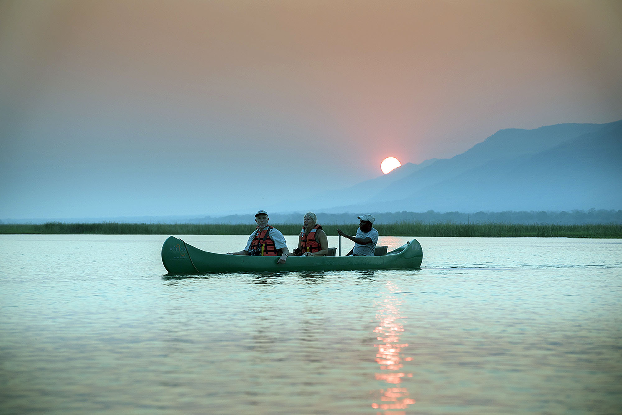 Gäste genießen eine Kanufahrt auf einem ruhigen See bei Sonnenuntergang vor einer Bergkulisse in Afrika.
